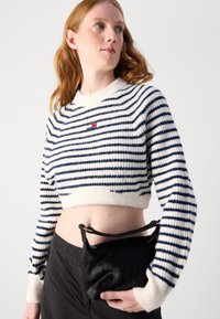 Stripet cropped genser i hvit og marineblå med ribbestrikket hals; paret med en liten svart veske med pelskvalitet som har en stropp.