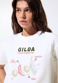 T-shirt en coton blanc présentant un design graphique coloré d'un piment, d'une olive et d'une anchois avec le texte "GILDA, un en-cas pour pimenter le tout."