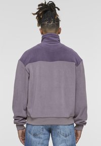 Sudadera de forro polar morada con cuello alto, que presenta un panel en un morado más claro en la parte superior de la espalda. Los puños y el dobladillo son ligeramente elásticos.