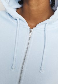 Ljusblå zip-up hoodie med en mjuk tygtextur, justerbara snörningar och en silvrig dragkedjedetalj framtill.