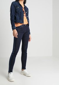 Person wearing dark blue denim jacket and jeans, orange graphic T-shirt, and white sneakers, standing against a plain light background.