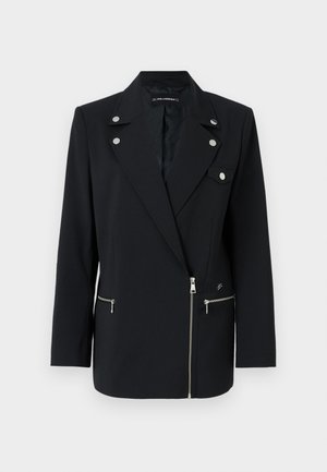 Musta räätälöity bleiseri, jossa on hopeanvärisiä yksityiskohtia, varustettu kauluksella, vetoketjullisilla etutaskuilla ja epäsymmetrisellä muotoilulla.