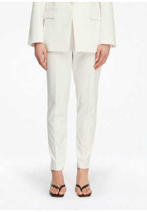 Kobieta w dopasowanych białych spodniach i pasującym białym żakiecie, w czarnych sandałach na obcasie z paskami, stojąca na białym tle.