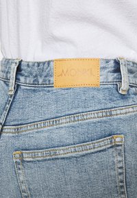 Vaqueros de mezclilla azul deslavado con costuras amarillas, que presentan una etiqueta de cuero marcada "MONKI" en la cintura y un detalle en el bolsillo trasero.