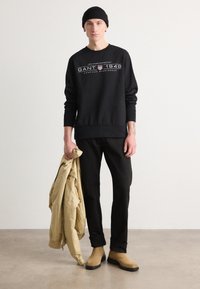 Svart hettegenser med hvit grafisk tekst, svarte bukser, beige jakke holdt i hånden, beige støvler, minimalistisk bakgrunn.