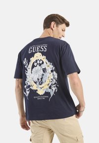 Camiseta de algodón azul marino con un estampado floral en gris y oro en la parte trasera, con el texto "GUESS" y "ROPA DE CALIDAD DESDE 1981".