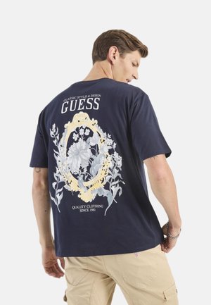 Camiseta de algodón azul marino con un estampado floral en gris y oro en la parte trasera, con el texto "GUESS" y "ROPA DE CALIDAD DESDE 1981".