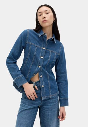 Joven mujer con chaqueta vaquera abotonada y jeans a juego, posando con una mano en la cintura sobre un fondo liso.