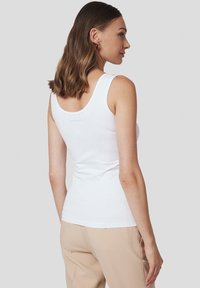 Biała bezrękawnik z okrągłym dekoltem z tyłu, wykonany z gładkiego materiału. Zawiera subtelny detal logo w okolicy dekoltu. Dopasowany krój.