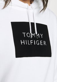 Felpa bianca con cappuccio caratterizzata da un grande rettangolo nero con la scritta "TOMMY HILFIGER" in lettere bianche, cordini in morbido tessuto.