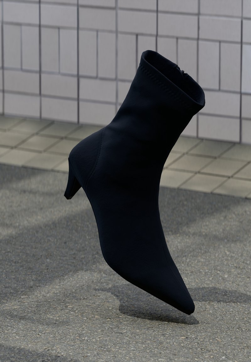 Bottine noire à bout pointu avec un petit talon, flottant au-dessus d'un trottoir pavé avec un mur carrelé en arrière-plan.