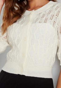 Krátký rukáv, krémový cardigan s pleteným vzorem má žebrovaný lem a tři knoflíky vzadu, což vytváří texturovaný efekt.