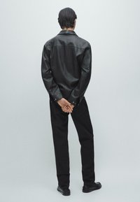 Sort læderjakke med krave, lange ærmer og knappede manchetter, kombineret med sorte bukser og sko. Skjorten har en glat tekstur.