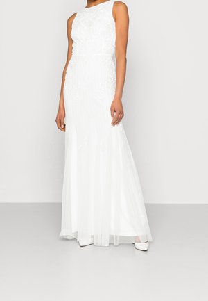 Woman wearing sleeveless white embroidered floor-length gown and white pointed-toe heels, standing against a plain light background.