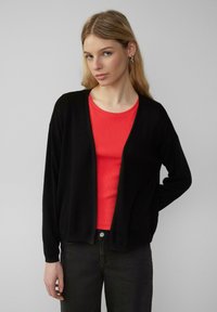 Cardigan noir en tissu doux, design à ouverture frontale, manches longues, associé à un t-shirt rouge à côtes et un jean foncé. Style minimaliste.