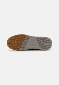 Semelle d'une chaussure décontractée avec une bande de roulement en caoutchouc segmentée marron et grise présentant des motifs d’adhérence texturés et un détail du nom de la marque au centre.