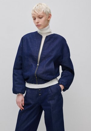 Persoon met kort blond haar draagt een marineblauwe jas met rits over een witte coltrui en bijpassende marineblauwe broek, staand tegen een grijze achtergrond.