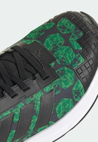 Czarny but sportowy z teksturowaną, wzorzystą górą zdobioną zielonymi wzorami w kształcie kostek, czarnym systemem sznurowania oraz białą podeszwą.