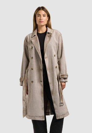 Woman wearing a beige double-breasted trench coat over a black top and black pants, standing against a plain light gray background.