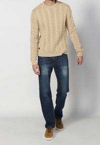 Pull beige en maille torsadée avec poignets côtelés, associé à un jean en denim bleu. Des chaussures en daim marron soulignent un look décontracté.