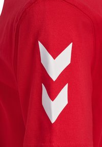 Rød bomulds-T-shirt med en detalje ved venstre skulder, der har to hvide chevron-striber, som fremhæver en glat tekstur og en tætsiddende pasform.