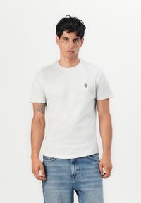Hellgraues Baumwoll-T-Shirt mit kurzen Ärmeln, Rundhalsausschnitt und kleinem schwarzen Wappensymbol auf der Brust, kombiniert mit blauen Jeans.