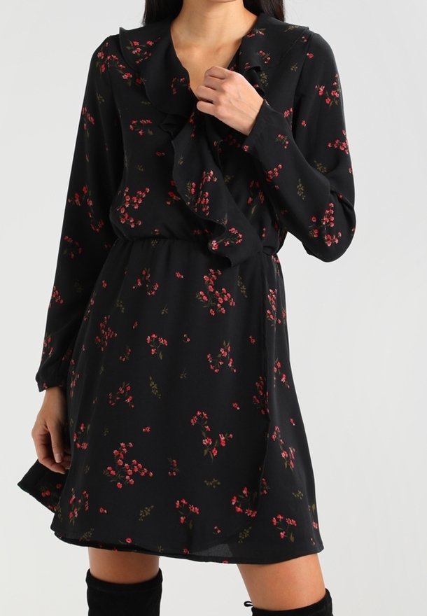 Femme portant une robe noire à manches longues avec des motifs floraux rouges et un col volanté, associée à des bottes noires cuissardes.