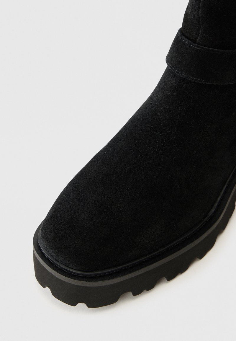 Botín de ante negro con punta redondeada, superficie texturizada y suela de goma negra gruesa que presenta un patrón de zigzag para mayor tracción.