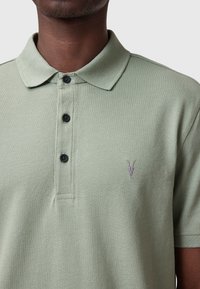 Pikétröja i ljusgrön bomull med en texturerad yta, tre svarta knappar och en subtil grå logotypdetalj på vänster bröst.