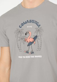 Grå bomulds-t-shirt med en tegneserieflamingo i solbriller, der holder et surfbræt, omgivet af palmer og tekst.