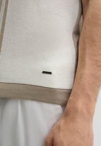 White mesh knit pullover with beige trim at the hem. Features a small black label with brand name near the bottom seam.