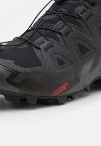 Chaussure de trail noire robuste avec embout texturé renforcé, panneaux en mesh, boucles pour lacets, et logo rouge « contagrip » sur le bord de la semelle.