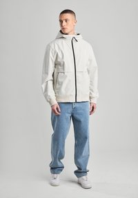 Giacca a cappuccio beige chiaro con zip nera, polsini elastici e un fit rilassato. Abbinata a jeans azzurri larghi e sneakers bianche.