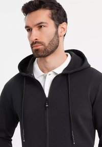 Sort zip-up hoodie med justerbar snorhætte, blød tekstur og kontrasterende hvid polo-krave under. Lynlåsen har sort hardware.