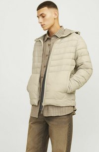 Veste matelassée beige clair avec des coutures horizontales, fermeture éclair et capuche. Portée par-dessus une chemise à carreaux et associée à un pantalon marron.