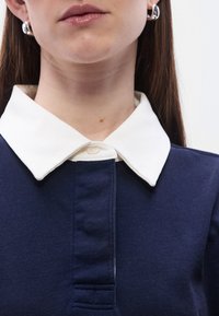 Πολο μπλε ναυτικού χρώματος με λευκό γιακά, με πλακέτα με κουμπιά και υφασμάτινη υφή. Λείο φινίρισμα, κοντά μανίκια και ορατές ραφές.