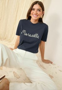 Tamsiai mėlyna marškinėliai su balta siuvinėta užrašu "Marseille", derinami su baltais aukšto liemens kelnėmis. Tekstūruota fone suteikia kontrasto.