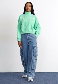 Felpa verde menta a zip con logo giallo, abbinata a pantaloni cargo in denim blu a vestibilità ampia con molte tasche e orlo sfrangiato.
