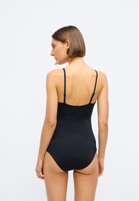 Femeie cu părul scurt și brunet, purtând un costum de baie negru cu bretele subțiri și ajustabile, văzută din spate pe un fundal simplu.