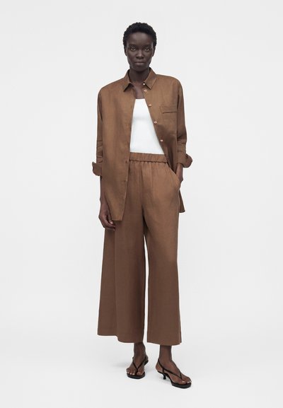 Ψηλό άτομο φοράει φαρδιά καφέ παντελόνια και ταιριαστή μακρυμάνικη μπλούζα πάνω από ένα λευκό αμάνικο τοπ και μαύρα σανδάλια με τακούνι, όρθιο.