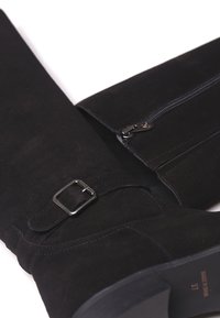 Las botas altas de ante negro cuentan con un zipper lateral, una correa ajustable con una hebilla rectangular y una textura mate suave.