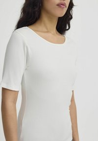 Femme aux cheveux bouclés et foncés portant un haut blanc côtelé à manches courtes et col en U, sur un fond clair uni.