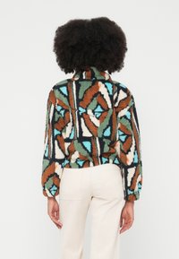 Flísová bunda s farebným geometrickým vzorom v zelenej, oranžovej, modrej a čiernej farbe. Má vysoký golier a dlhé rukávy.