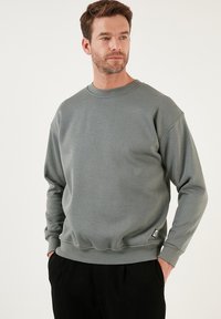 Sudadera gris de cuello redondo fabricada en una suave mezcla de algodón. Presenta puños y dobladillo acanalados, con una pequeña etiqueta de logo en la parte inferior izquierda.
