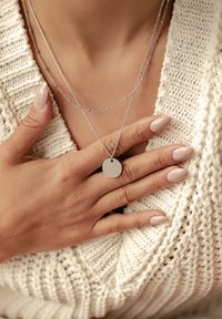 Collana d'argento con due catene che presenta un pendente piatto e circolare. Lo sfondo include un maglione lavorato a maglia bianco e unghie in tonalità nude curate.