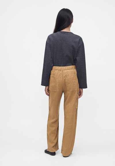Femme aux longs cheveux noirs portant une chemise à manches longues rayée noir et gris et un pantalon ample beige avec des poches appliquées, debout de dos.