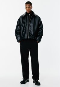 Veste en simili cuir noir avec grand col, fermeture éclair à l'avant et manches froncées, associée à un pantalon noir et des chaussures à lacets.