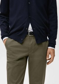 Cardigan a maglia blu navy con bottoni, abbinato a pantaloni verde oliva. Camicia bianca sotto, fissata con una cintura nera, che mostra una texture liscia.