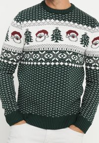Grüner gestrickter Pullover mit weißen Schneeflocken, Weihnachtsmann-Gesichtern und Weihnachtsbäumen in einem gemusterten Design. Gerippte Bündchen und Halsausschnitt.