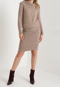 Pull en tricot beige avec un large col plié, associé à une jupe crayon ajustée. Des bottines en daim bordeaux complètent le look.